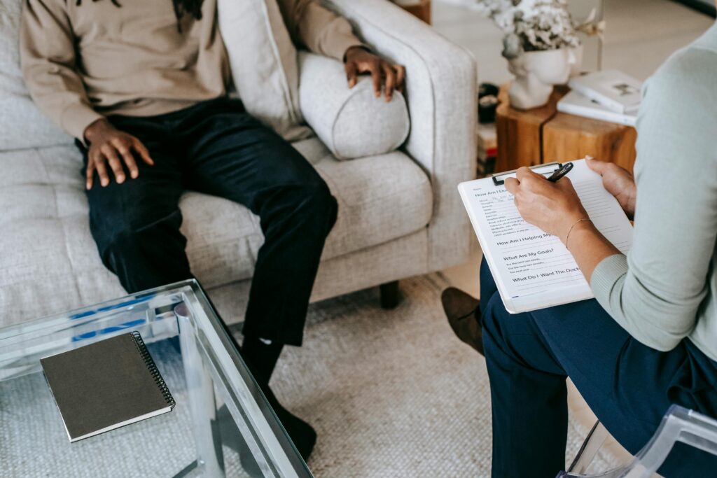 Valutazione e riabilitazione dei deficit cognitivi From above crop African American male in casual clothes sitting on comfy couch and answering professional female consultant questions in modern psychotherapy office